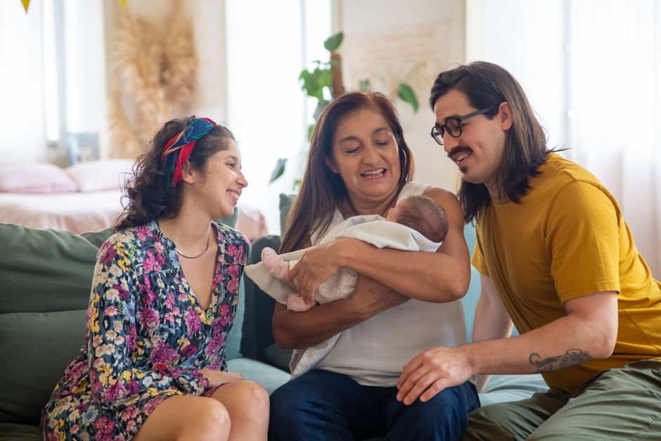 A grandmother happily holds her newborn grandchild while the baby's parents look on, representing the long-awaited arrival of a baby in the family.