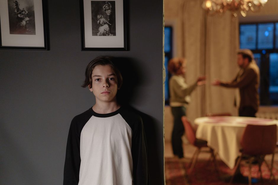 A teenager looks troubled while his parents argue in the background, illustrating the burden of sharing marital conflict with adult children.