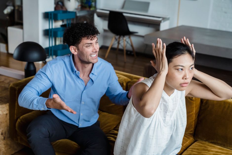 A woman shuts out her partner's demands, covering her ears as he argues on the couch.