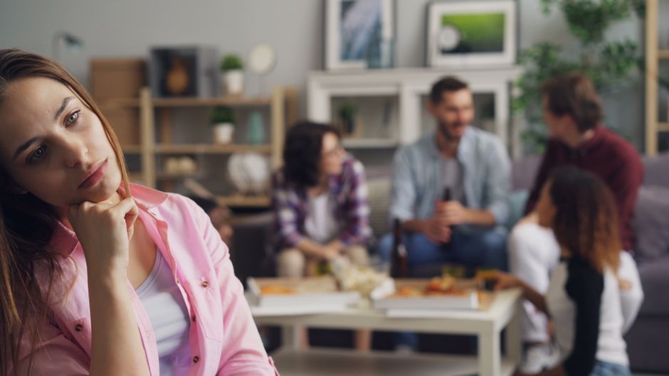 A woman feels excluded and alone while her friends gather together without her, reflecting the isolation of being kept apart from loved ones.