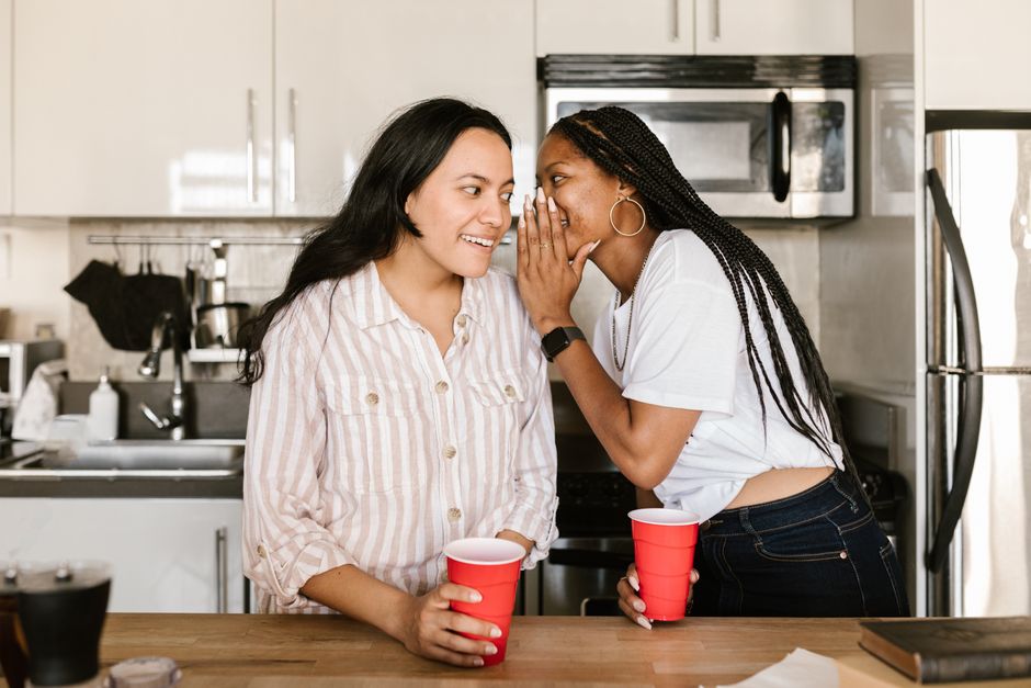 A woman whispers a secret into her friend's ear while they share drinks in the kitchen, hinting at gossip not meant to be trusted.