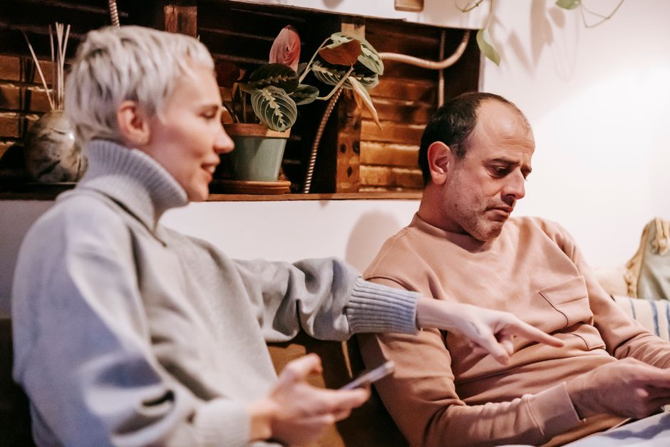 A man looks away distractedly as his partner tries to get his attention, reflecting the quiet tension of unspoken frustration in a relationship.