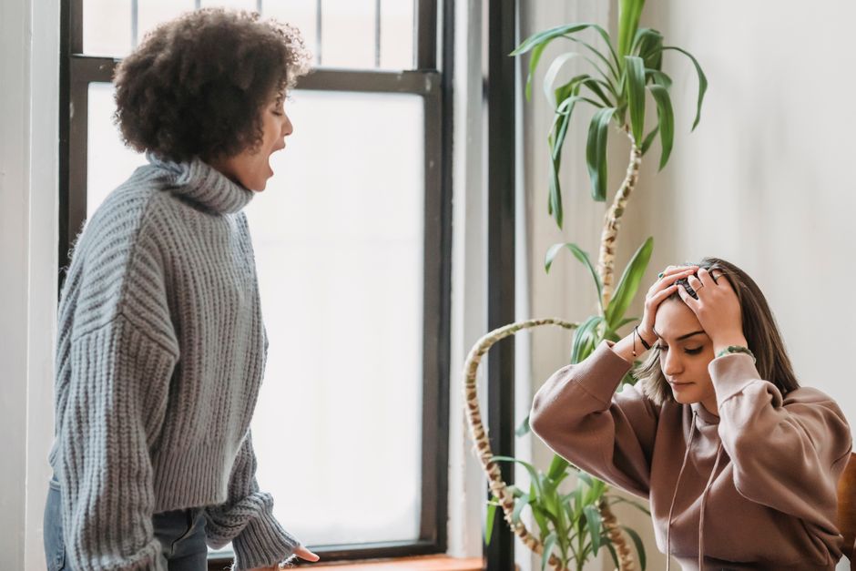 A woman reacts with visible stress, holding her head, while another woman yells at her, illustrating how reactions get blamed instead of the words spoken.