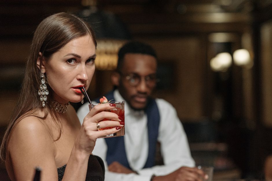 A woman lingers at the bar, sipping her drink slowly while casting a glance back, hoping someone will approach.