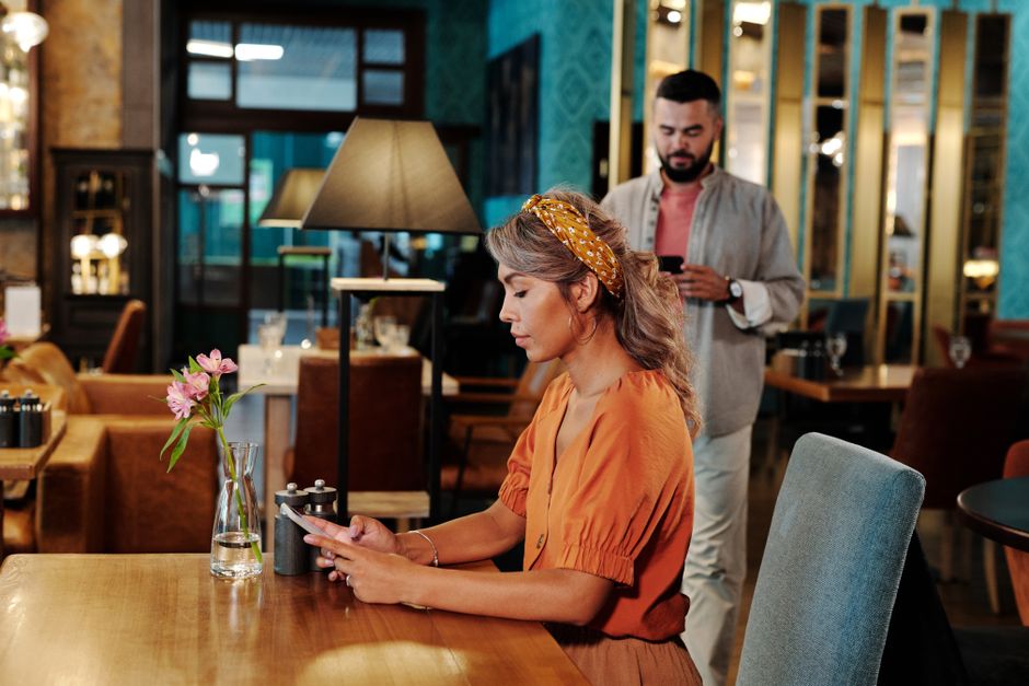 Woman checking her phone while a man walks up behind her at a café table.