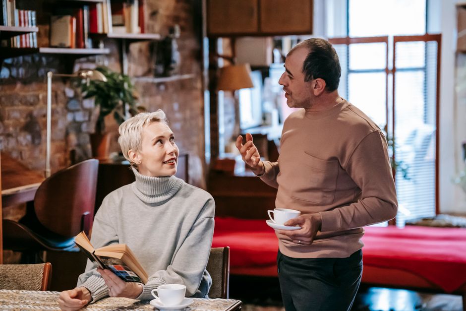 A couple in a heated discussion, with the man gesturing as if explaining himself and the woman watching skeptically, hinting at a conversation that doesn't quite add up.