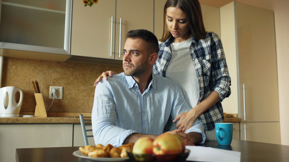 A woman offers quiet, steady comfort to a man struggling with a difficult moment, her hands resting supportively on his shoulders.